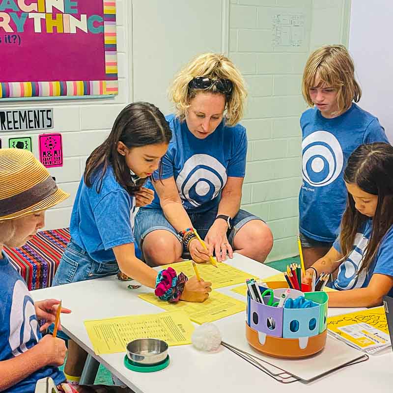 Fourth grade math teacher sits at table with micro school students to work on math problem.