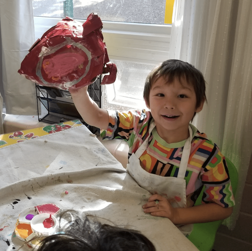 Private kindergarten student proudly raises paper mache creation.