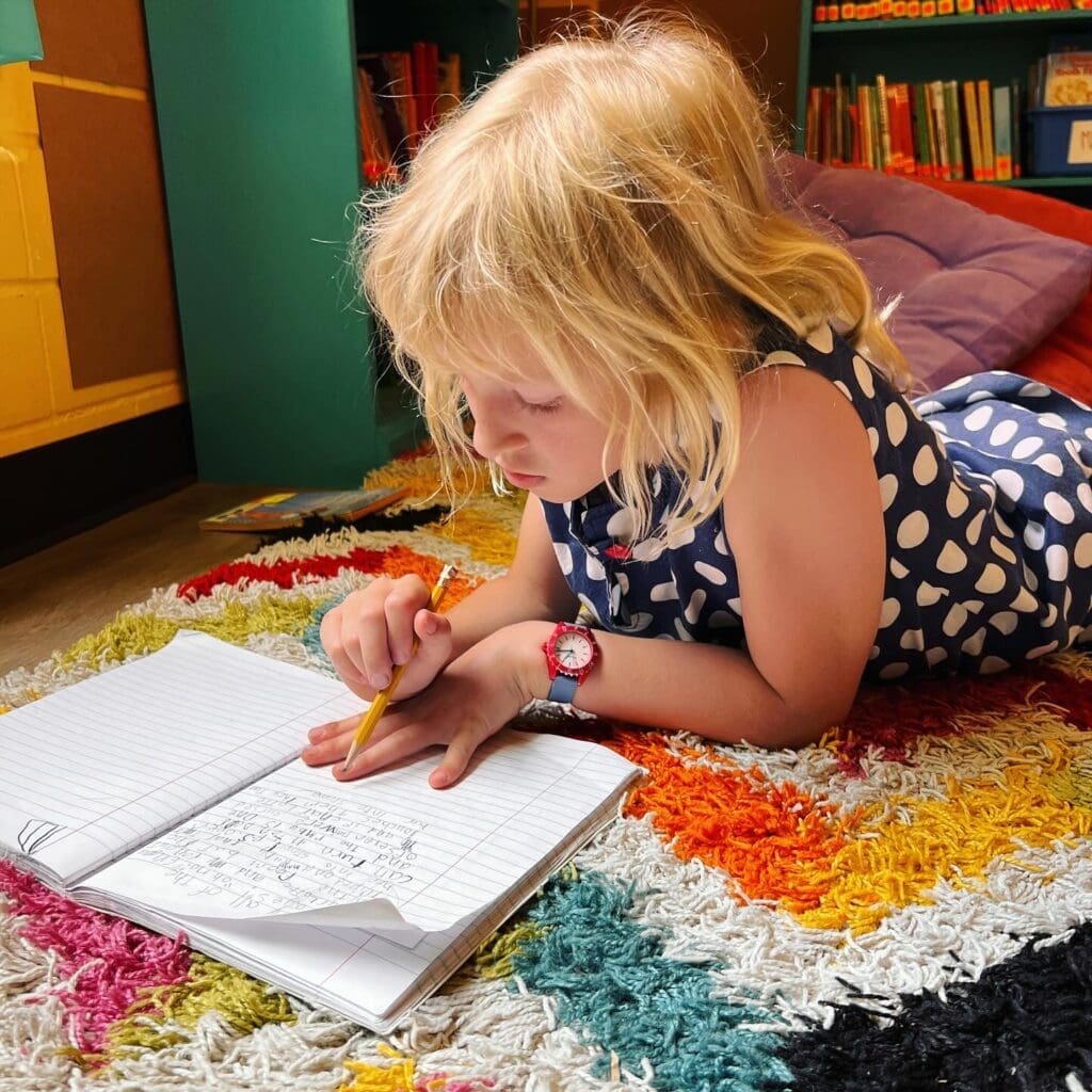 Second grade student writes while laying on rug at progressive school.